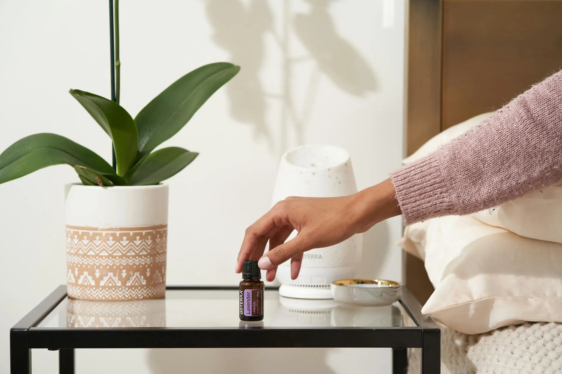 Daily scents that soothe the soul and calm the space Hand placing essential oil near diffuser on side table.